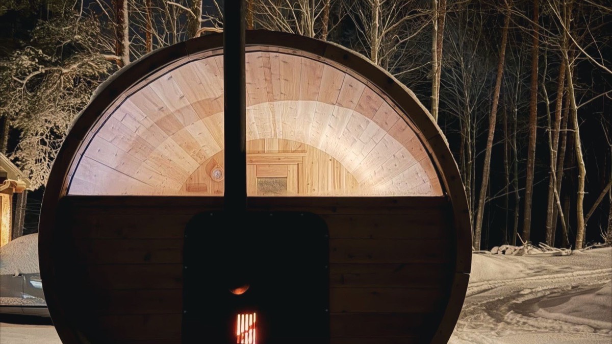 A wood-fired barrel sauna at night in the snow, lit warm from inside, with the stove door glowing.