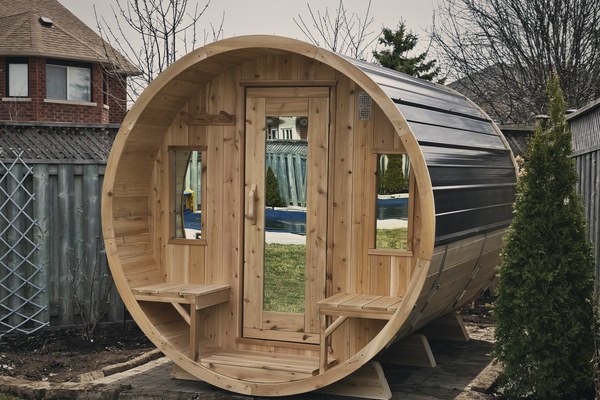 A cedar barrel sauna with a covered porch installed on a patio in a suburban backyard, beside a fence and a cedar hedge.