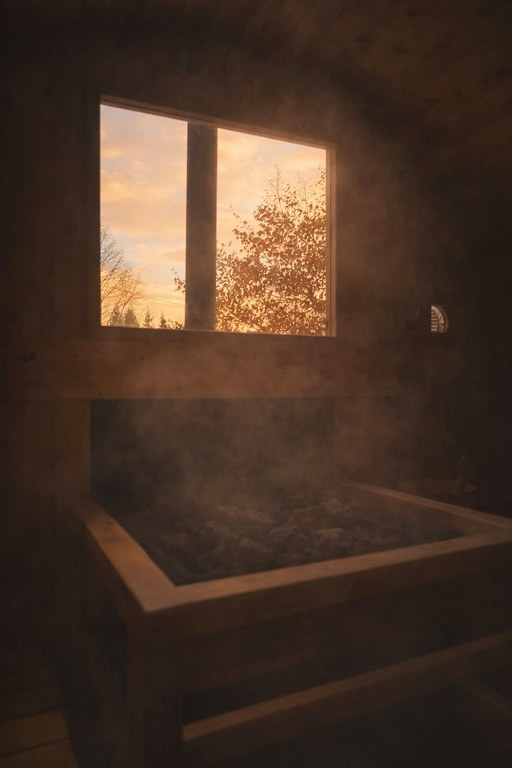 Inside the sauna at sunset — steam over the stove rocks, cedar benches, and a window looking out to bare trees.